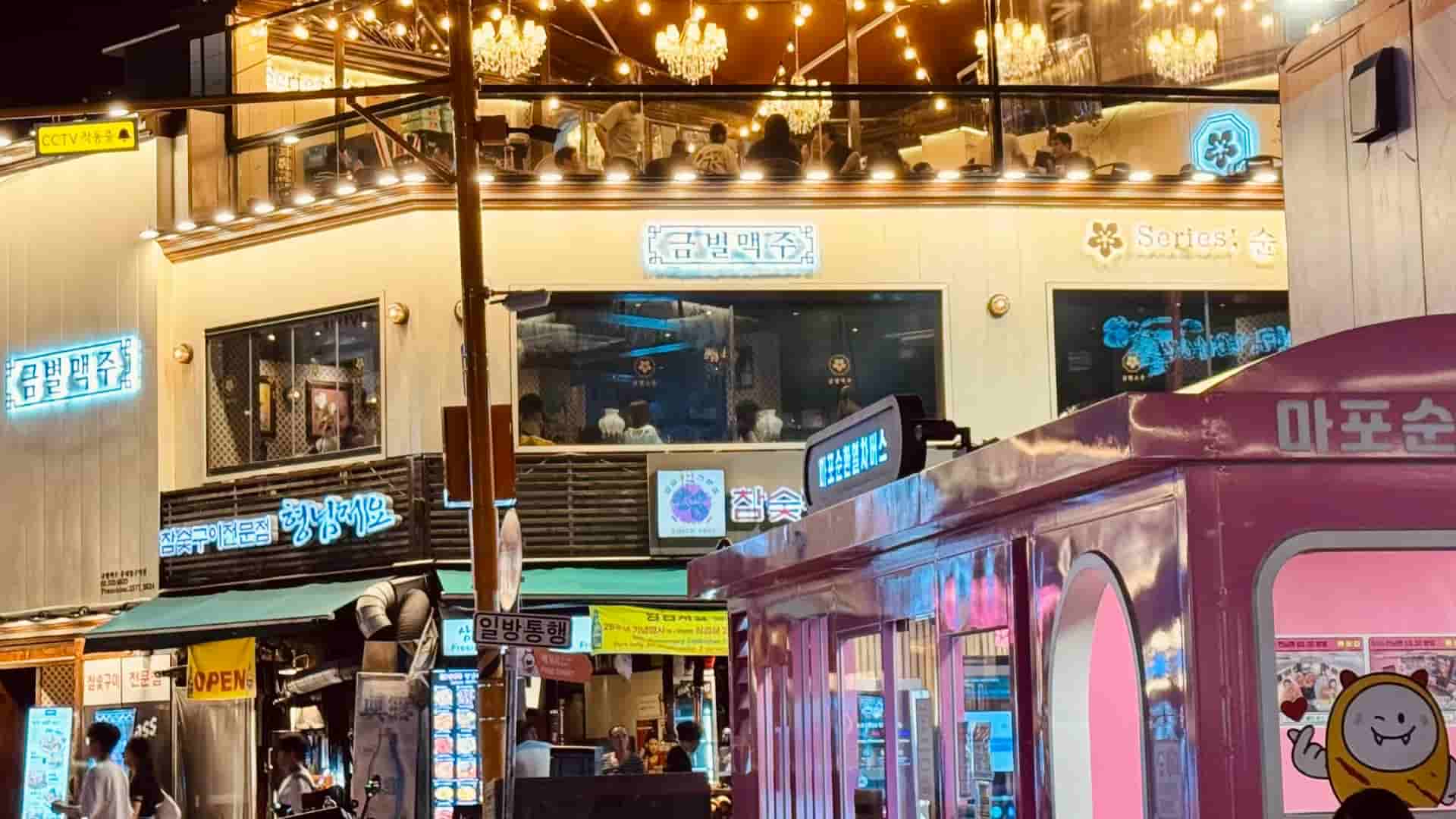 A vibrant multi-story building in Seoul at night, featuring a brightly lit open-air terrace with string lights, Korean neon signs, and a large pink train-shaped structure on the street level