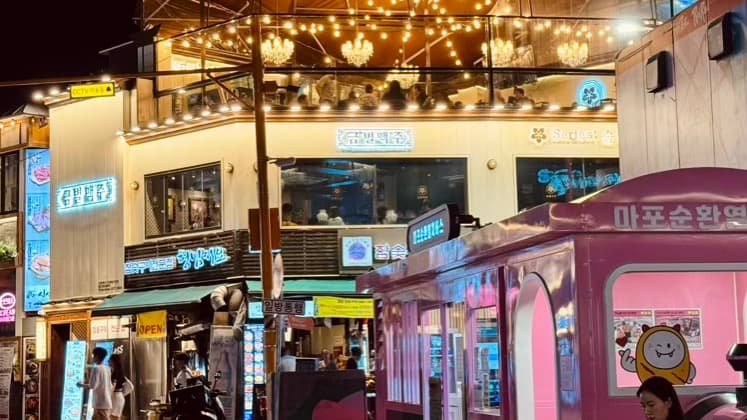 A vibrant multi-story building in Seoul at night, featuring a brightly lit open-air terrace with string lights, Korean neon signs, and a large pink train-shaped structure on the street level