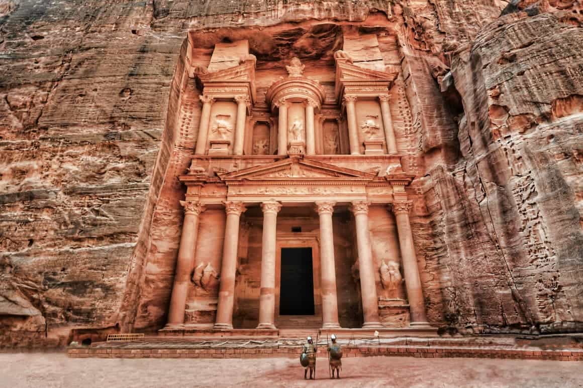 The iconic facade of The Treasury at Petra, Jordan, with two costumed guards standing in front of the massive entrance.