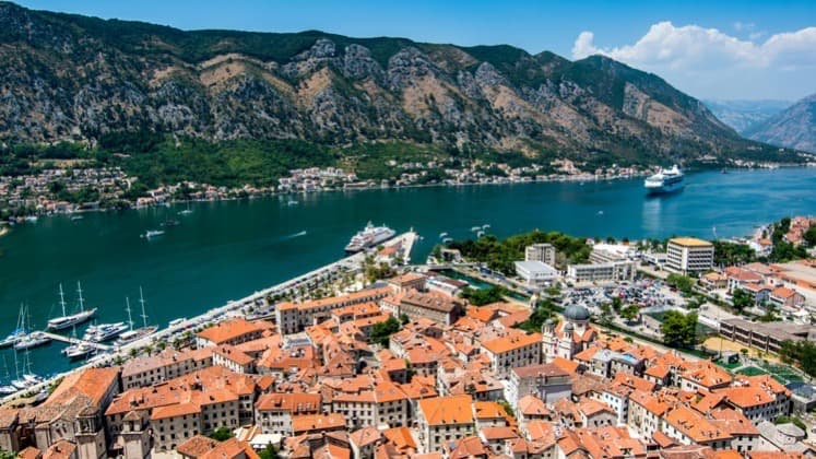 Aerial view of Kotor red tile roofs beside blue bay water and rugged mountains.