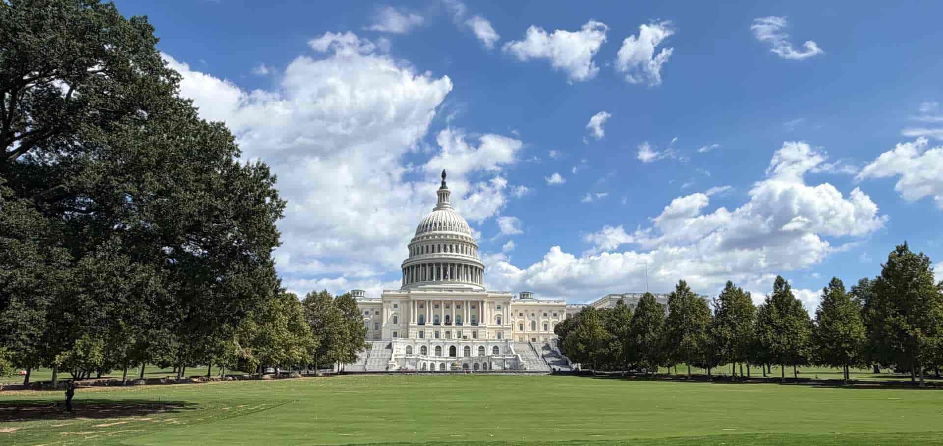 The iconic U.S. Capitol dome rising behind green lawns and leafy trees under a blue sky.