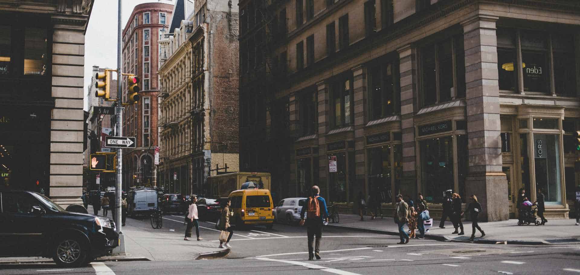 Pedestrians crossing a busy Manhattan street intersection framed by historic buildings and yellow cab