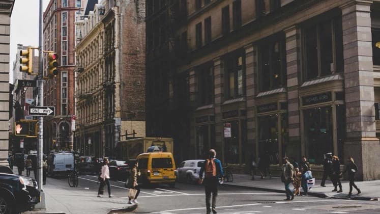 Pedestrians crossing a busy Manhattan street intersection framed by historic buildings and yellow cab