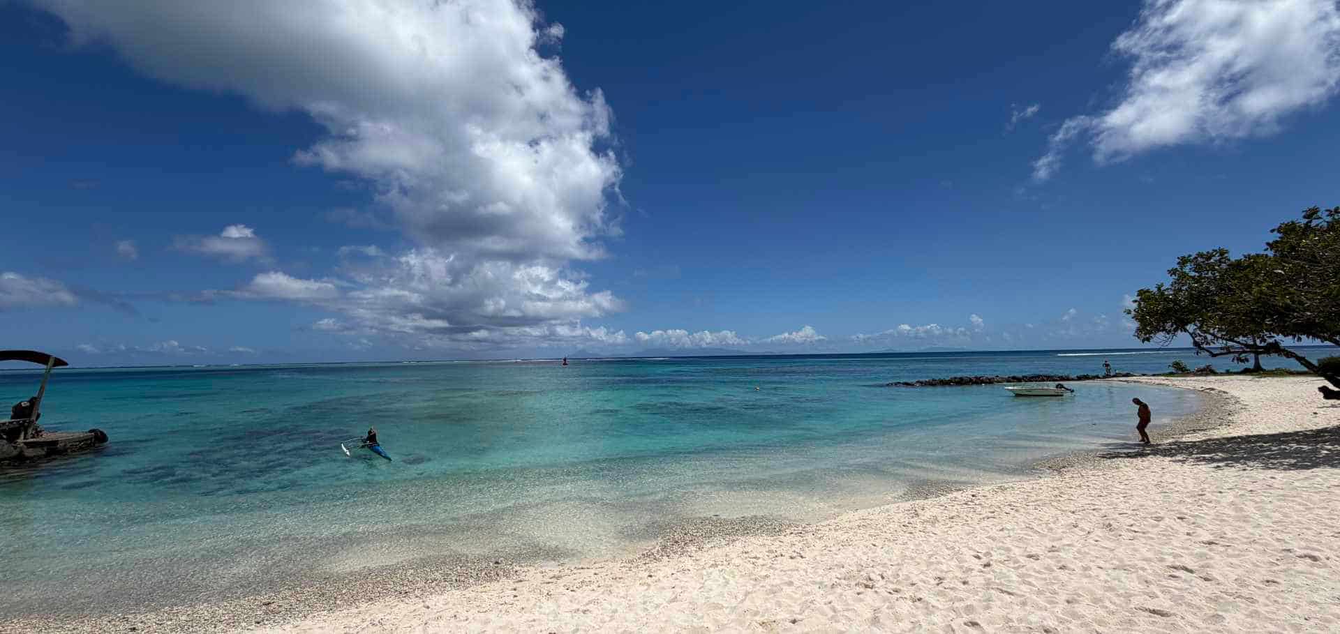 A person paddles a canoe along the turquoise shallows of a sandy beach in Huahine.