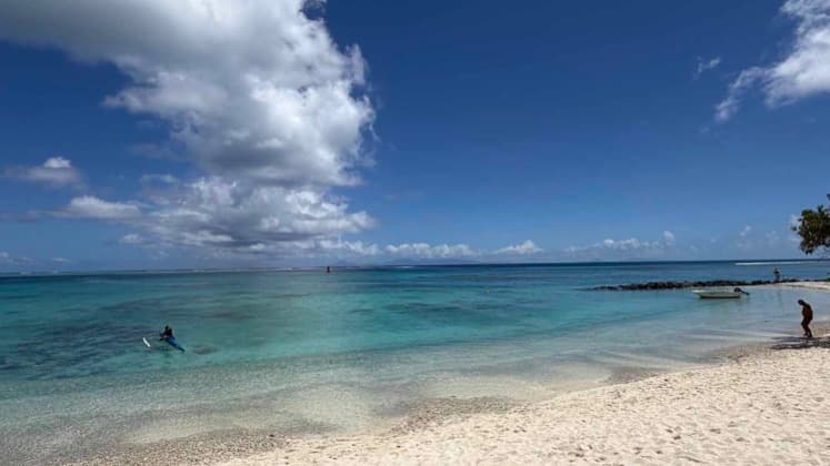 A person paddles a canoe along the turquoise shallows of a sandy beach in Huahine.
