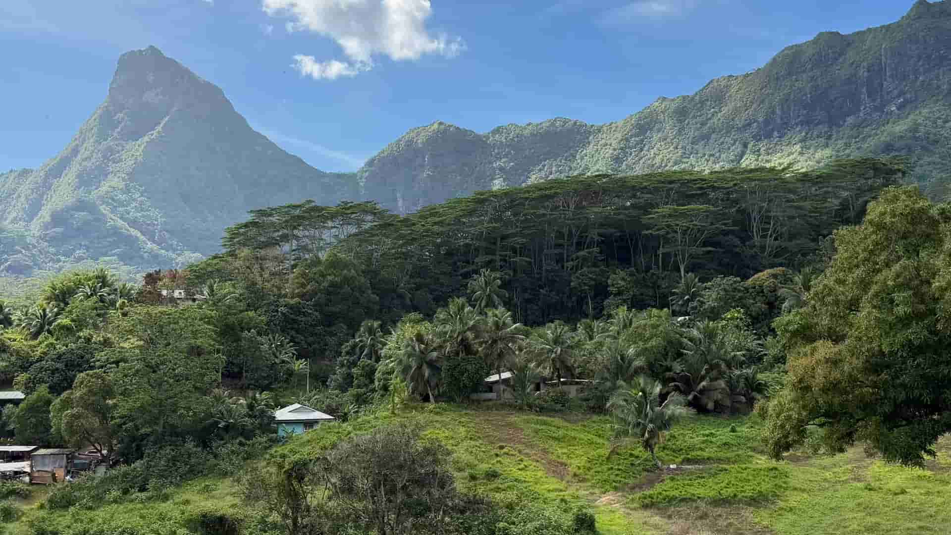 Lush green tropical valley with traditional houses nestled beneath towering volcanic mountain peaks.