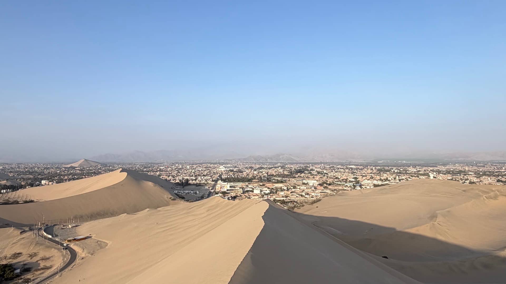 Aerial view of the lush Huacachina oasis village nestled among giant desert sand dunes under a clear blue sky.