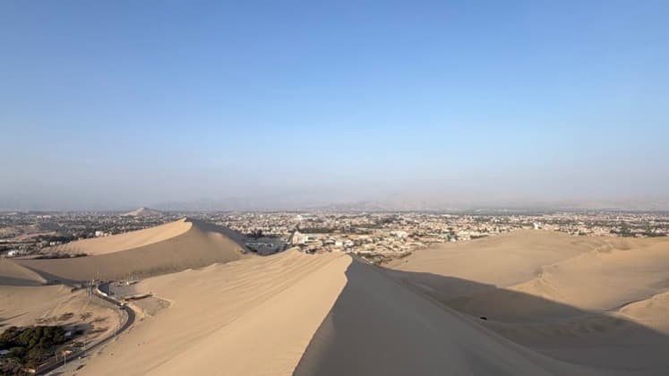 Aerial view of the lush Huacachina oasis village nestled among giant desert sand dunes under a clear blue sky.