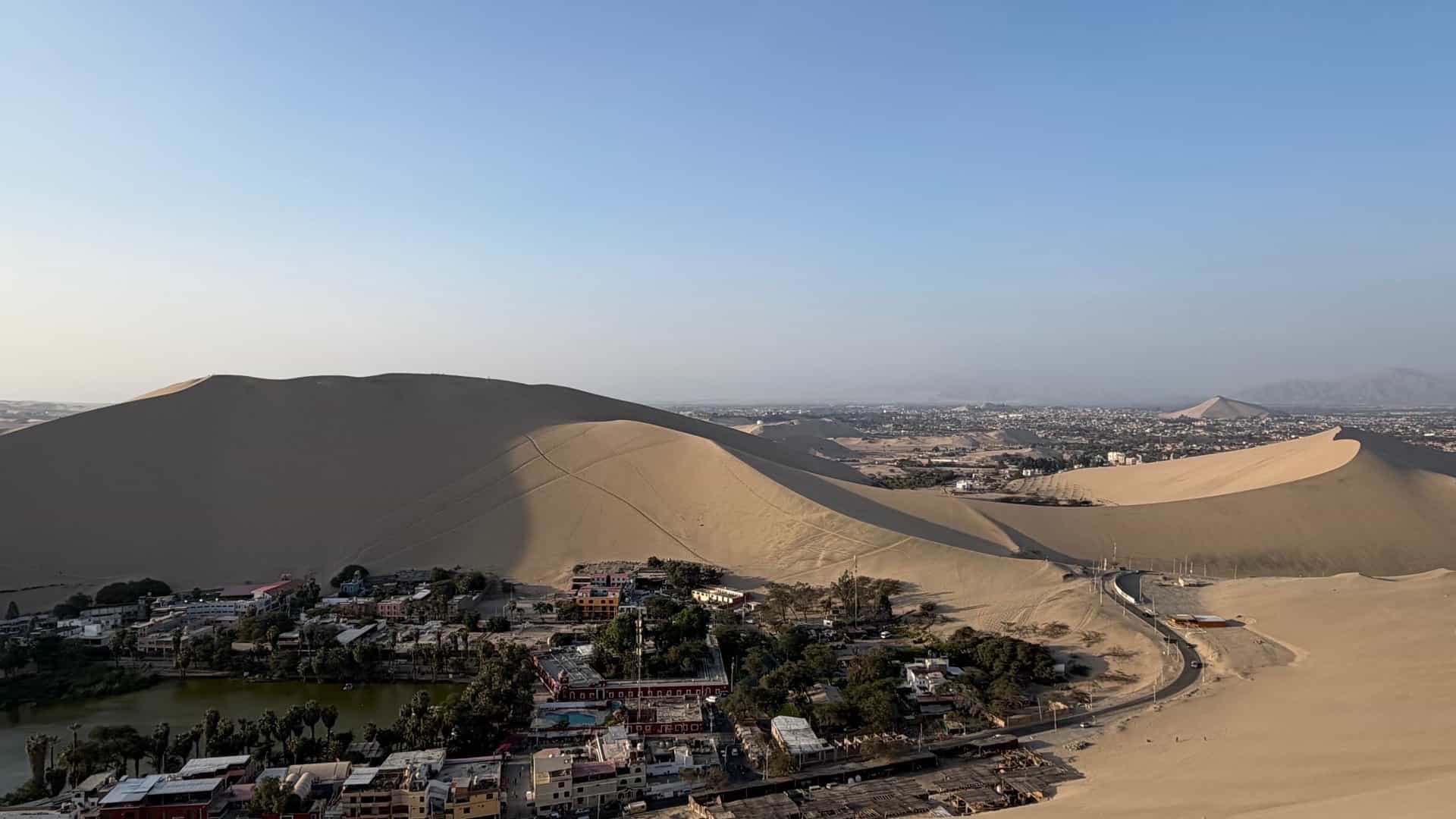 Aerial view of the lush green Huacachina oasis lagoon surrounded by giant sand dunes.