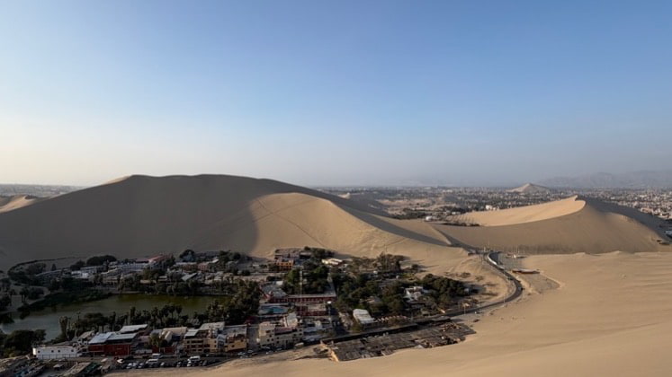 Aerial view of the lush green Huacachina oasis lagoon surrounded by giant sand dunes.
