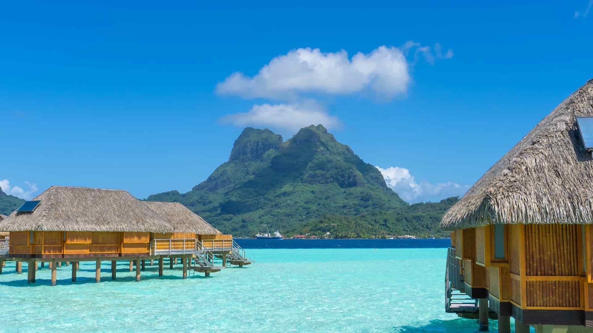 Overwater thatched bungalows standing above crystal-clear turquoise lagoon waters with green volcanic mountains.
