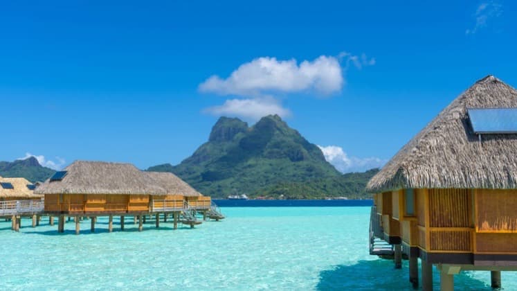 Overwater thatched bungalows standing above crystal-clear turquoise lagoon waters with green volcanic mountains.