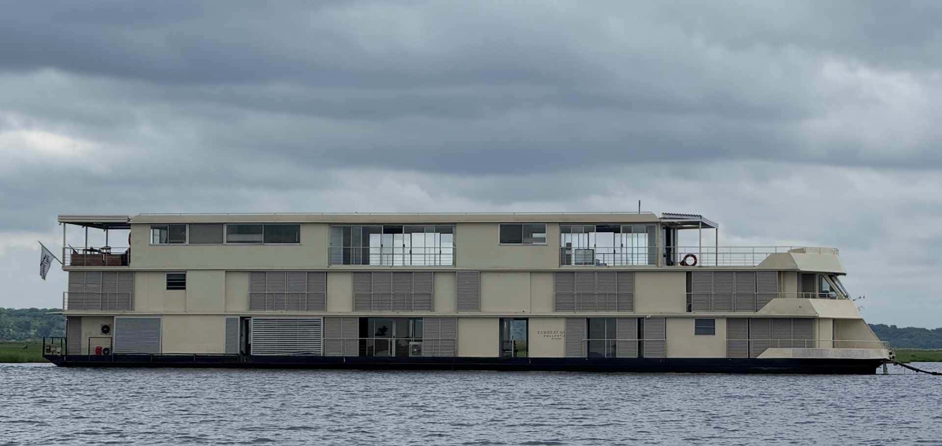 A large multi-story houseboat cruising along the calm waters of the Chobe River.