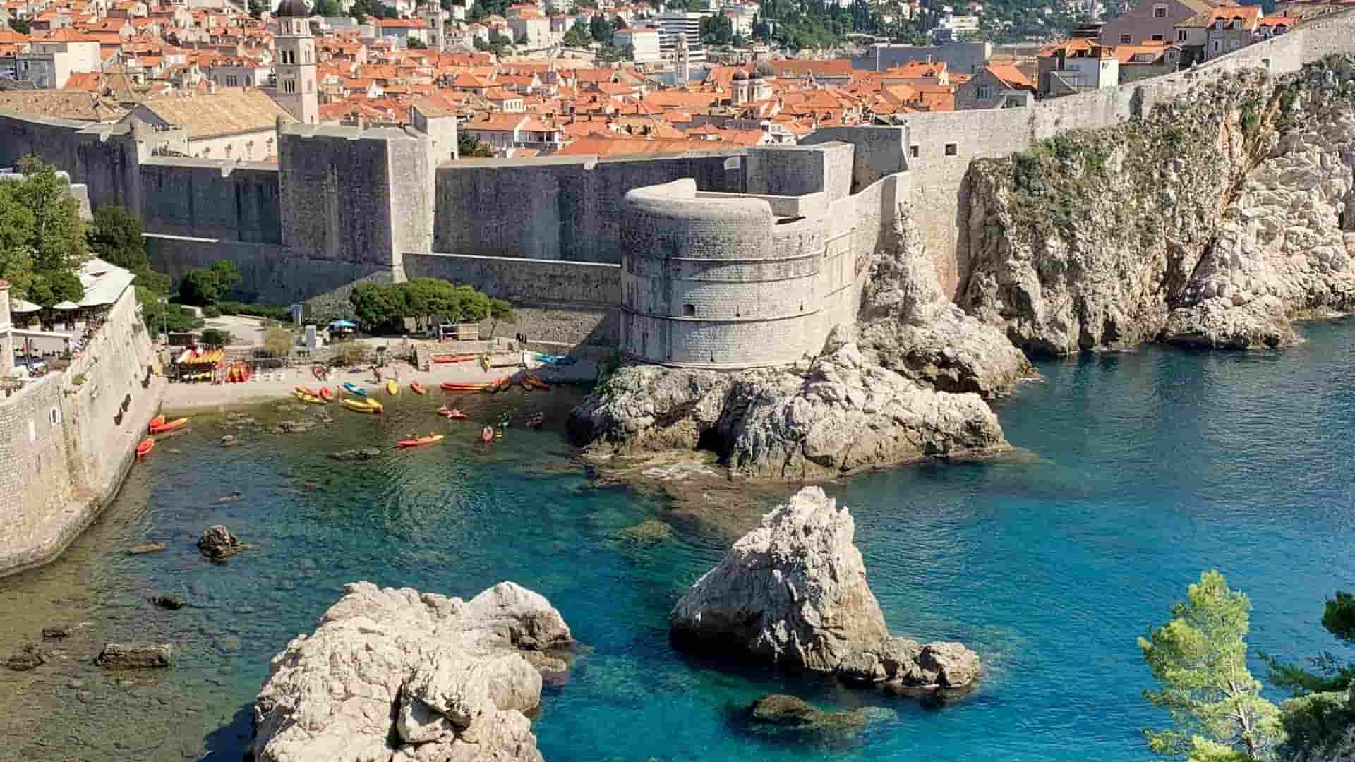 A view of the massive stone city walls of Dubrovnik meeting the turquoise Adriatic Sea, with a small beach below featuring colorful kayaks and rocky outcrops