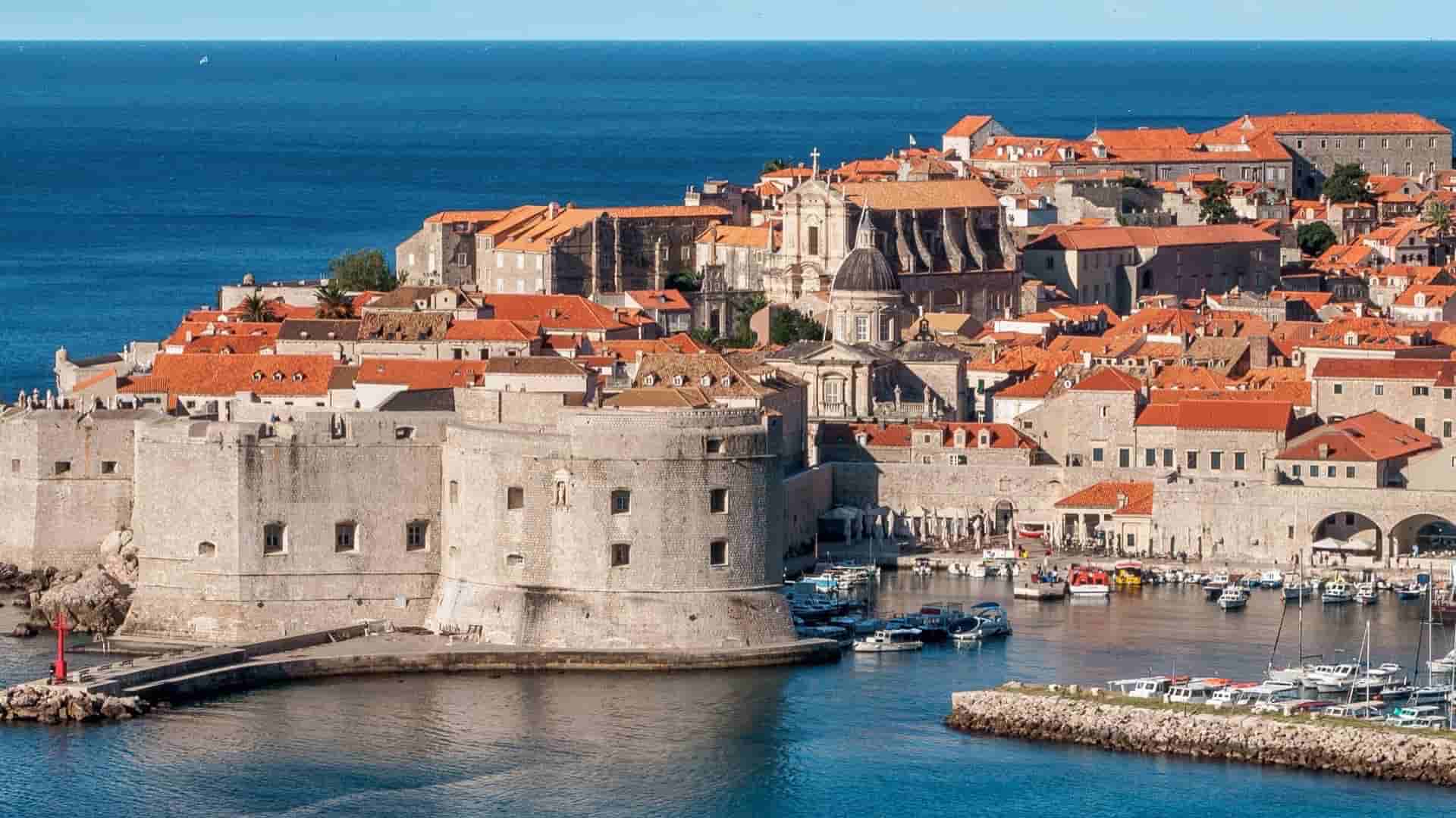 A daytime harbor view of Dubrovnik, highlighting the iconic orange-tiled roofs, historic stone fortifications, and moored boats resting in the calm blue water
