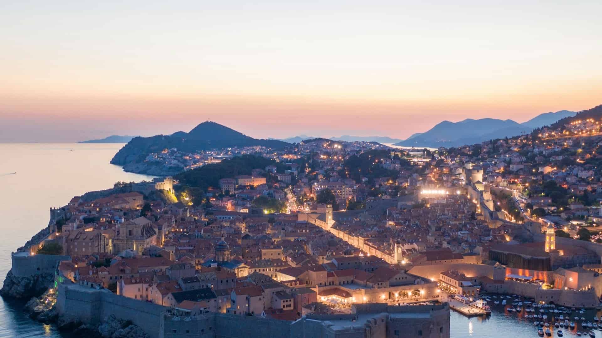An aerial view of the walled Dubrovnik Old Town at twilight, with warm lights illuminating the historic city streets and harbor against a soft sunset sky