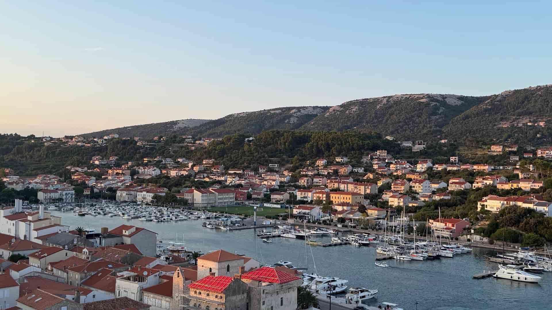 Elevated view of Rab harbor and marina filled with boats beneath green hills at sunset.