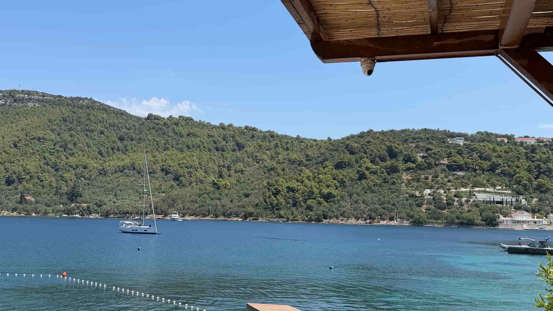 A white sailboat anchored in a calm, clear blue bay, viewed from behind a wooden pier with a shaded reed roof in the foreground, with lush green hills in the background