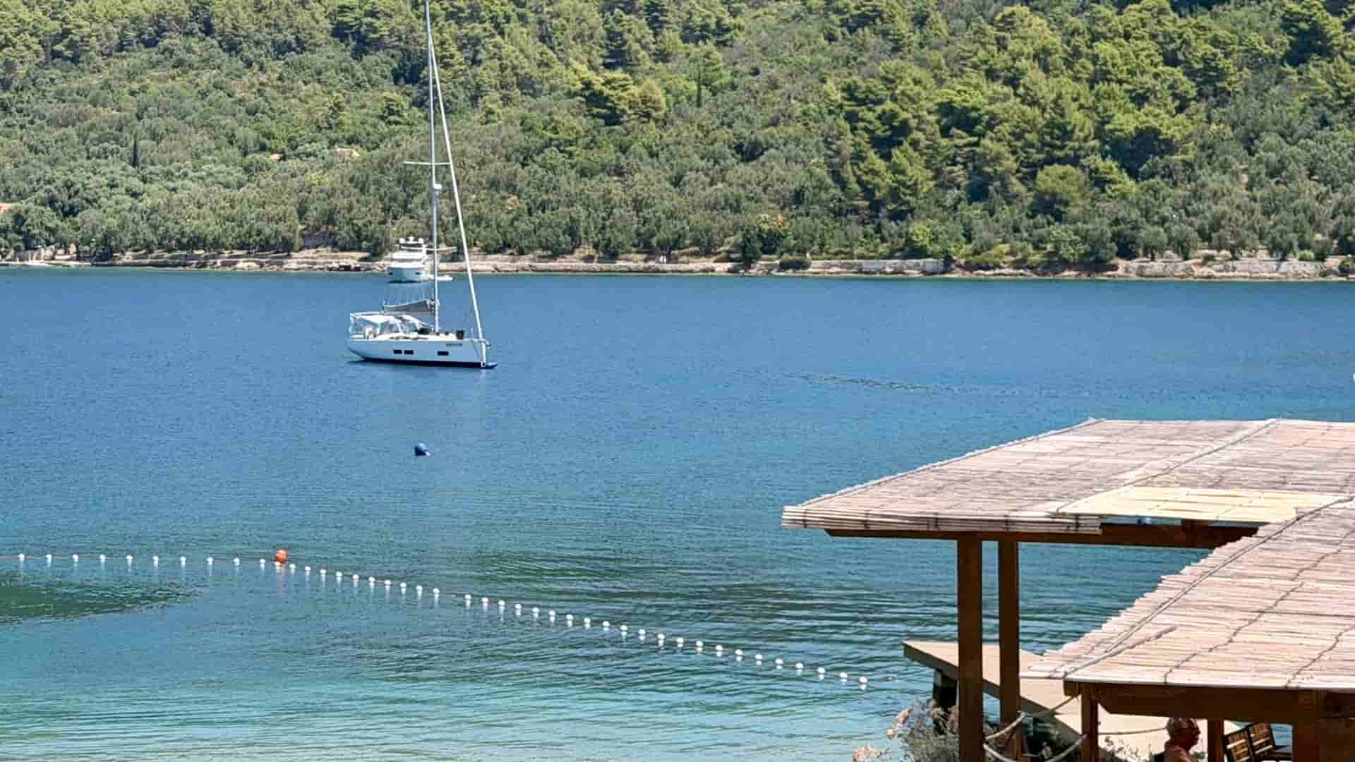 A wide, scenic view of a peaceful harbor with an anchored yacht, framed by the edge of a wooden canopy and surrounded by rolling hills covered in dense forest