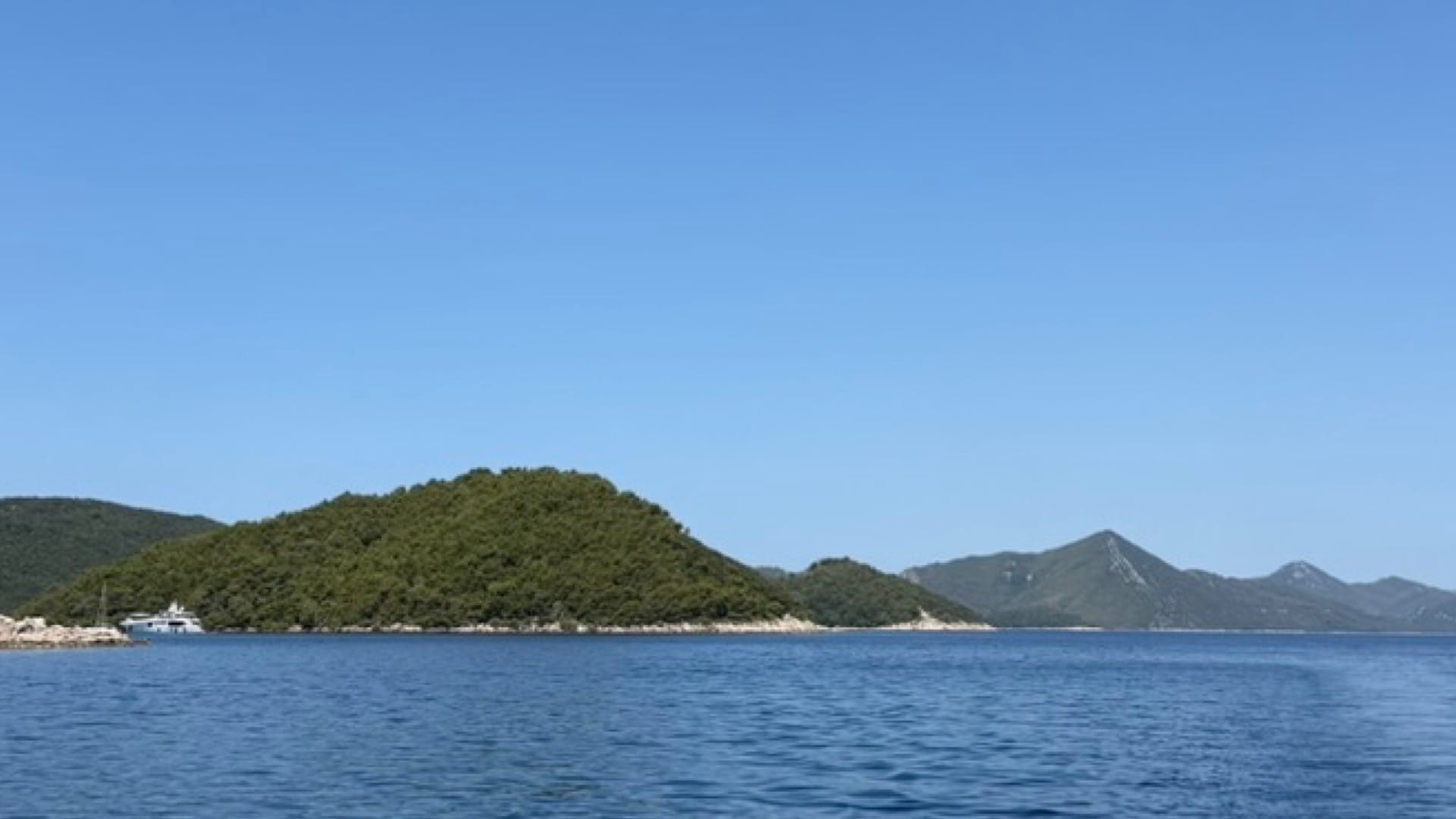 A wide, scenic view of a peaceful harbor with an anchored yacht, framed by the edge of a wooden canopy and surrounded by rolling hills covered in dense forest