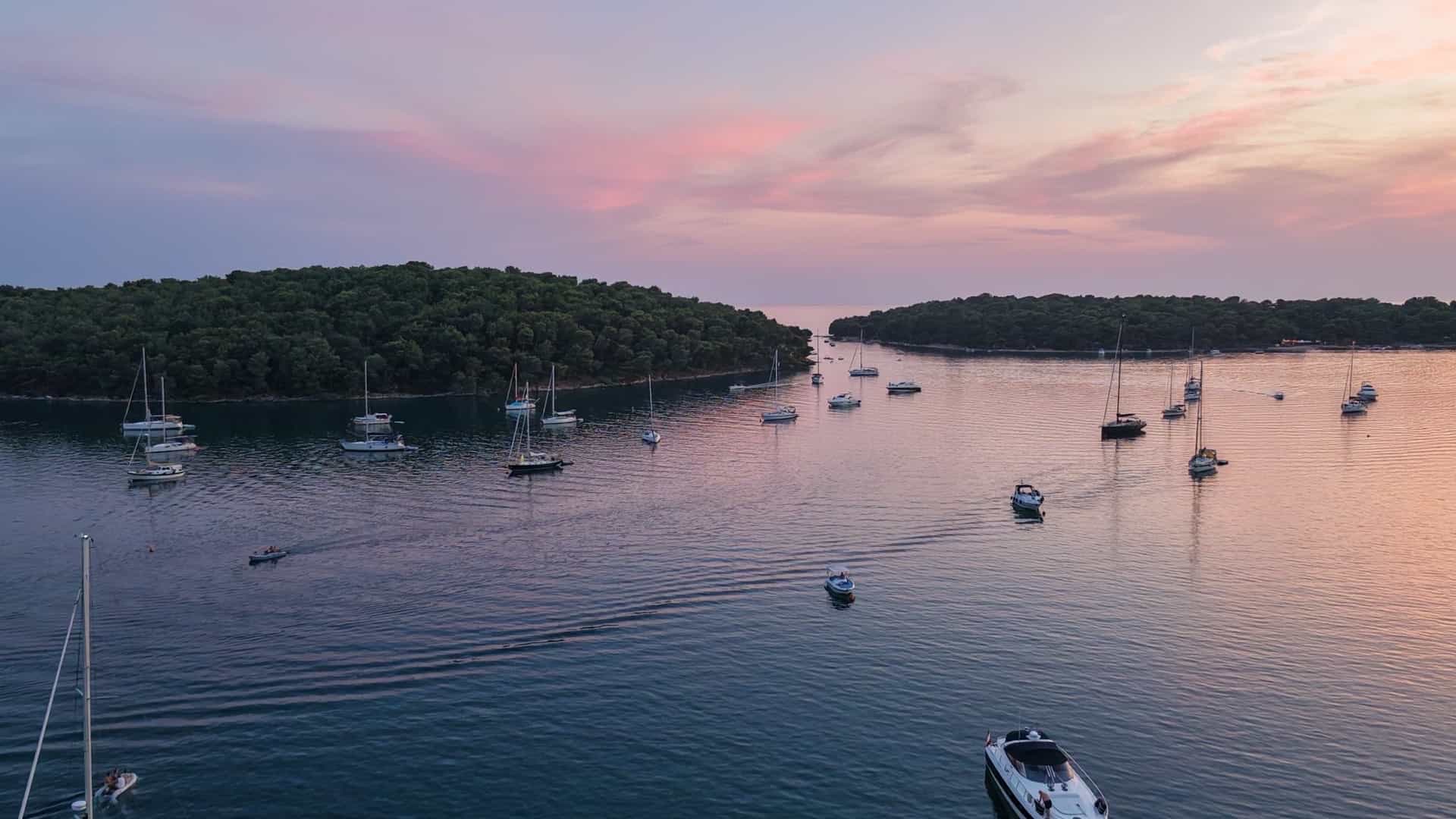 Sailboats navigating the pine-fringed bay channel of Uvala Soline during a pink sunset.