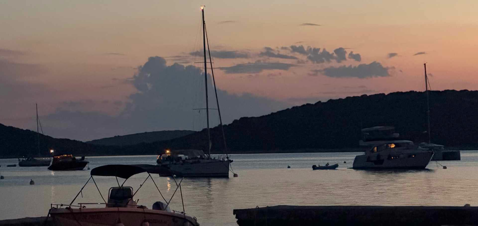 Calm evening anchorage with sailing yachts silhouetted against a colorful coastal dusk