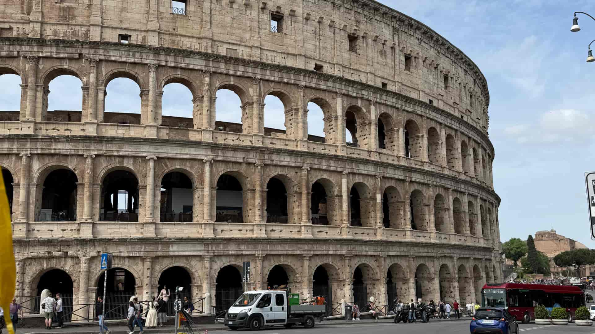 The massive stone arches of the Colosseum in Rome under a bright daytime sky with passing traffic.