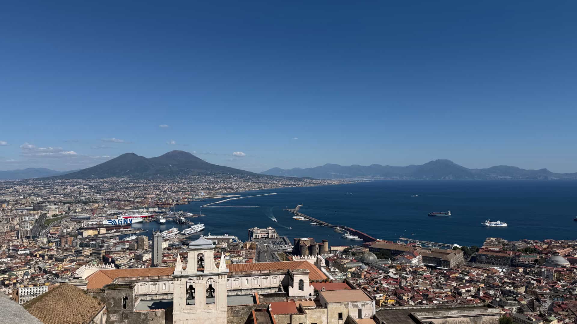 Panoramic view of Naples and Mount Vesuvius across the blue harbor from a high vantage point.