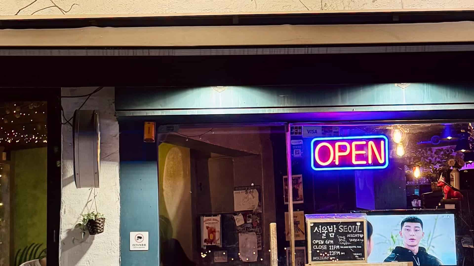 The exterior storefront of a bar at night, displaying a brightly glowing neon OPEN sign, a chalkboard reading "서울밤 SEOUL NIGHT", and a television screen visible through the glass.