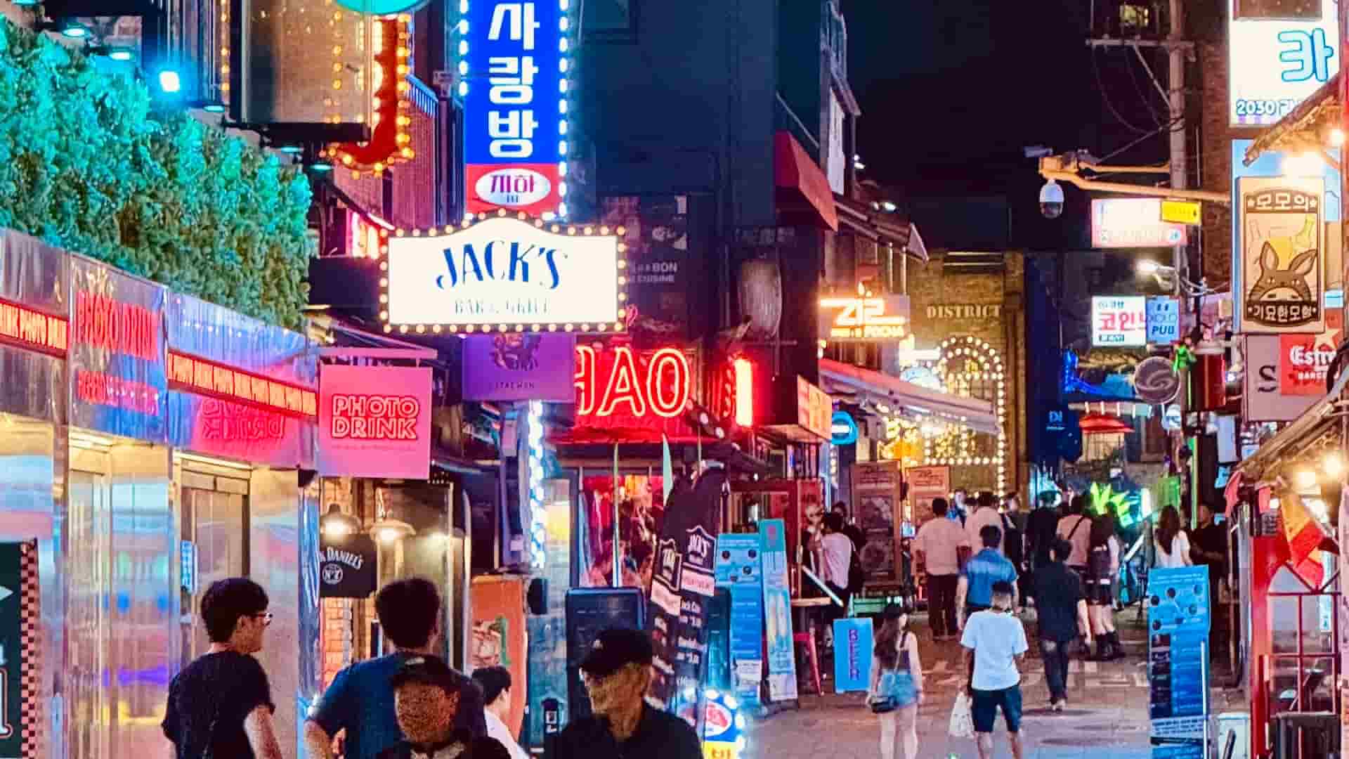 A bustling commercial street scene in Seoul at night, crowded with pedestrians walking past brightly illuminated storefronts, outdoor clothing racks, and assorted neon signs.