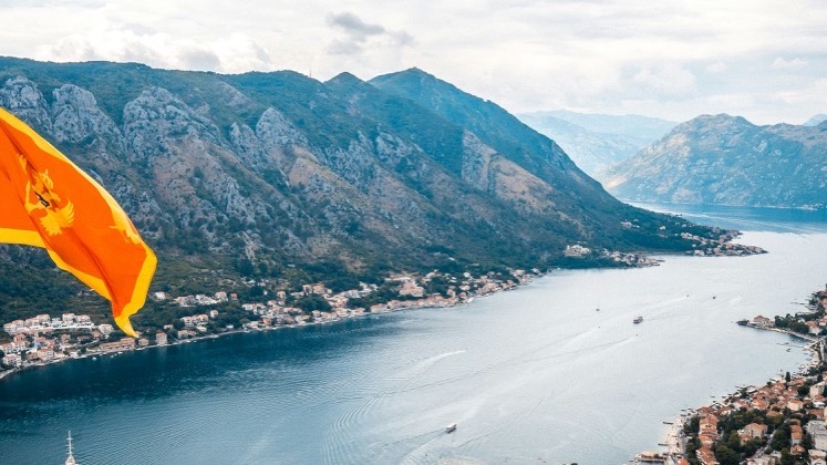 Montenegrin flag waving over a high-angle panoramic view of the Bay of Kotor.