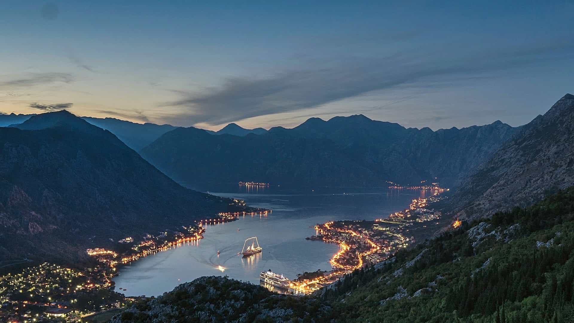 Illuminated shoreline along Bay of Kotor framed by steep dark mountains at dusk.