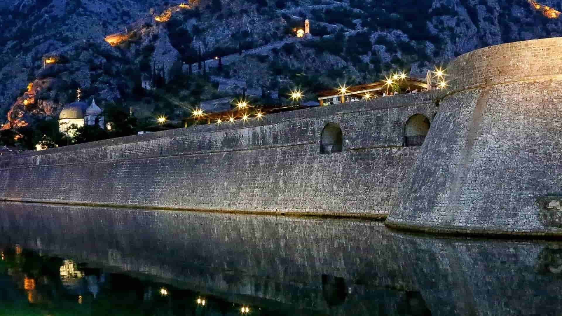 Floodlit stone fortress walls of Kotor reflecting in calm dark waters at night.