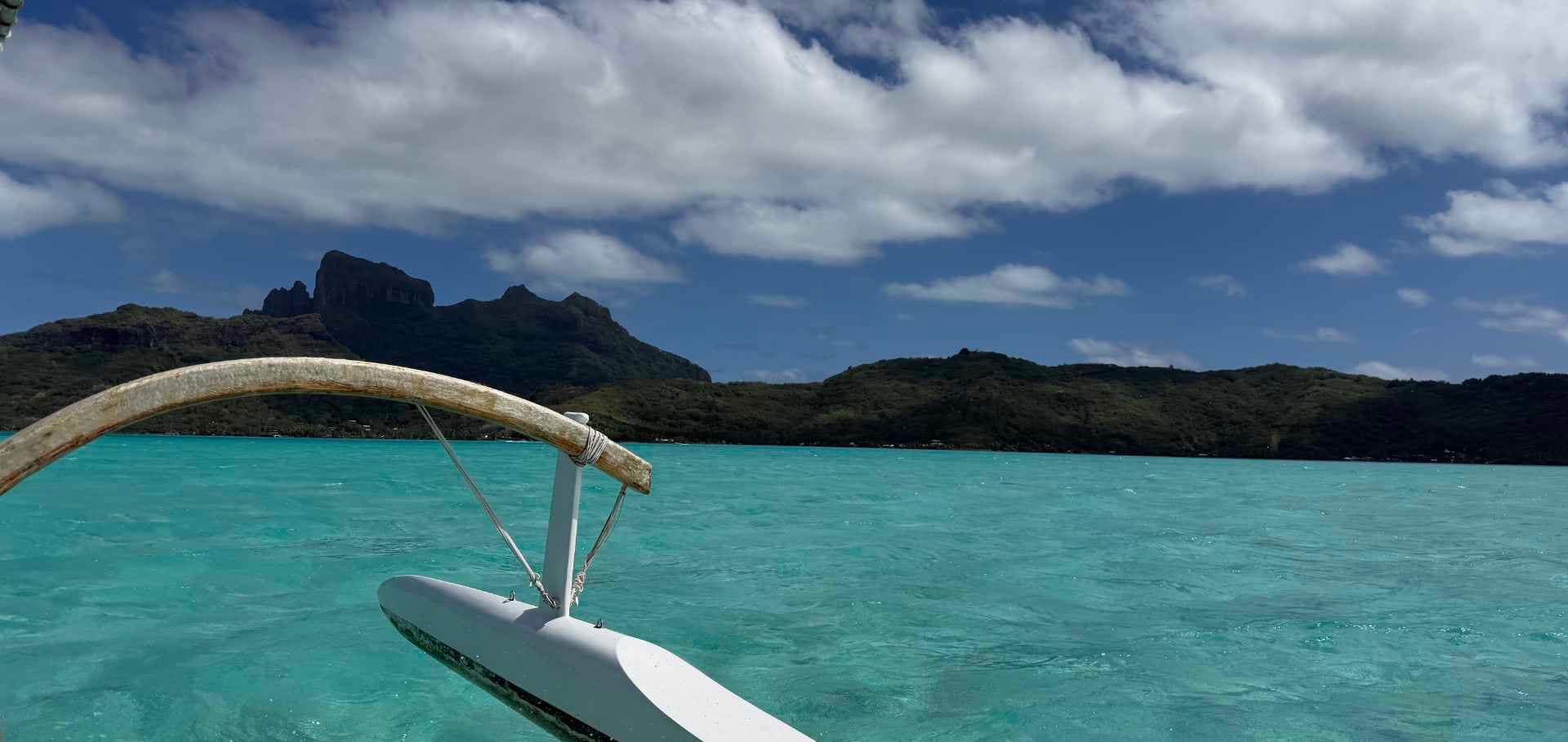 The traditional outrigger canoe prow cutting through shallow turquoise lagoon waters toward mountain peaks.