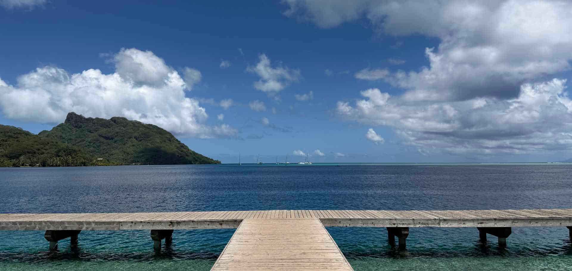 A wooden pier stretches out over the deep blue waters toward a lush volcanic island.