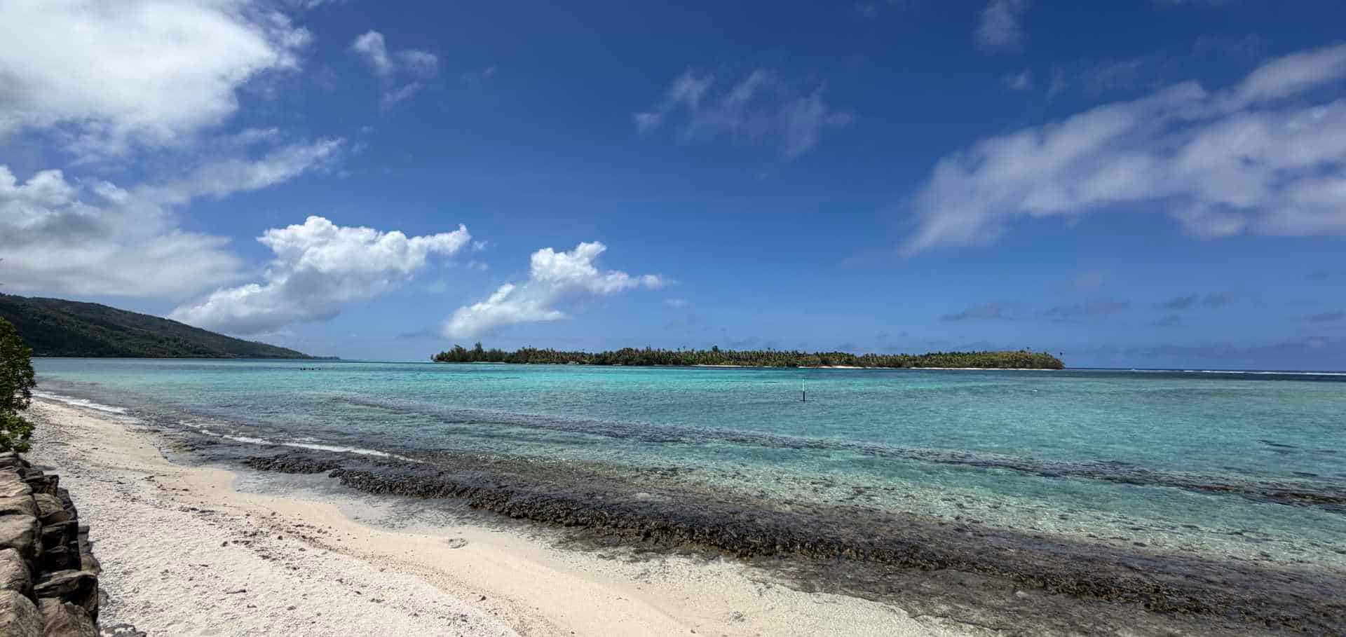 Gentle ocean waves wash onto a white sand beach with a distant tropical island under a blue sky.
