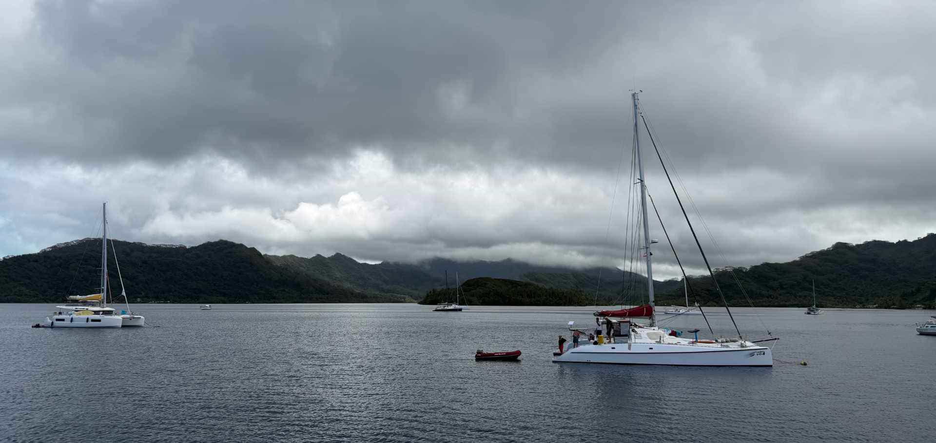 Sailboats anchored in a calm bay beneath overcast skies and mountainous terrain.