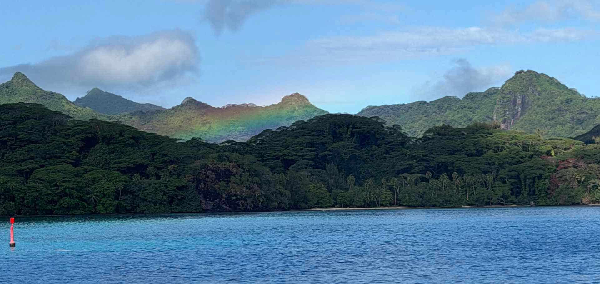 A faint rainbow arches over lush green mountain peaks under a partly cloudy sky.