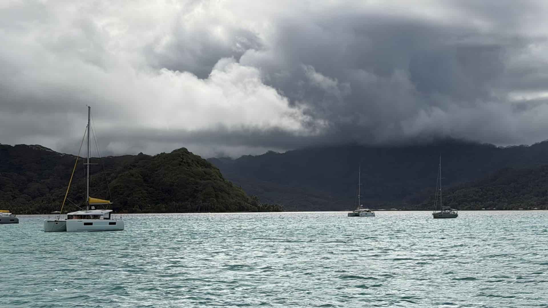 Sailboats anchored in a calm turquoise lagoon beneath dramatic storm clouds.