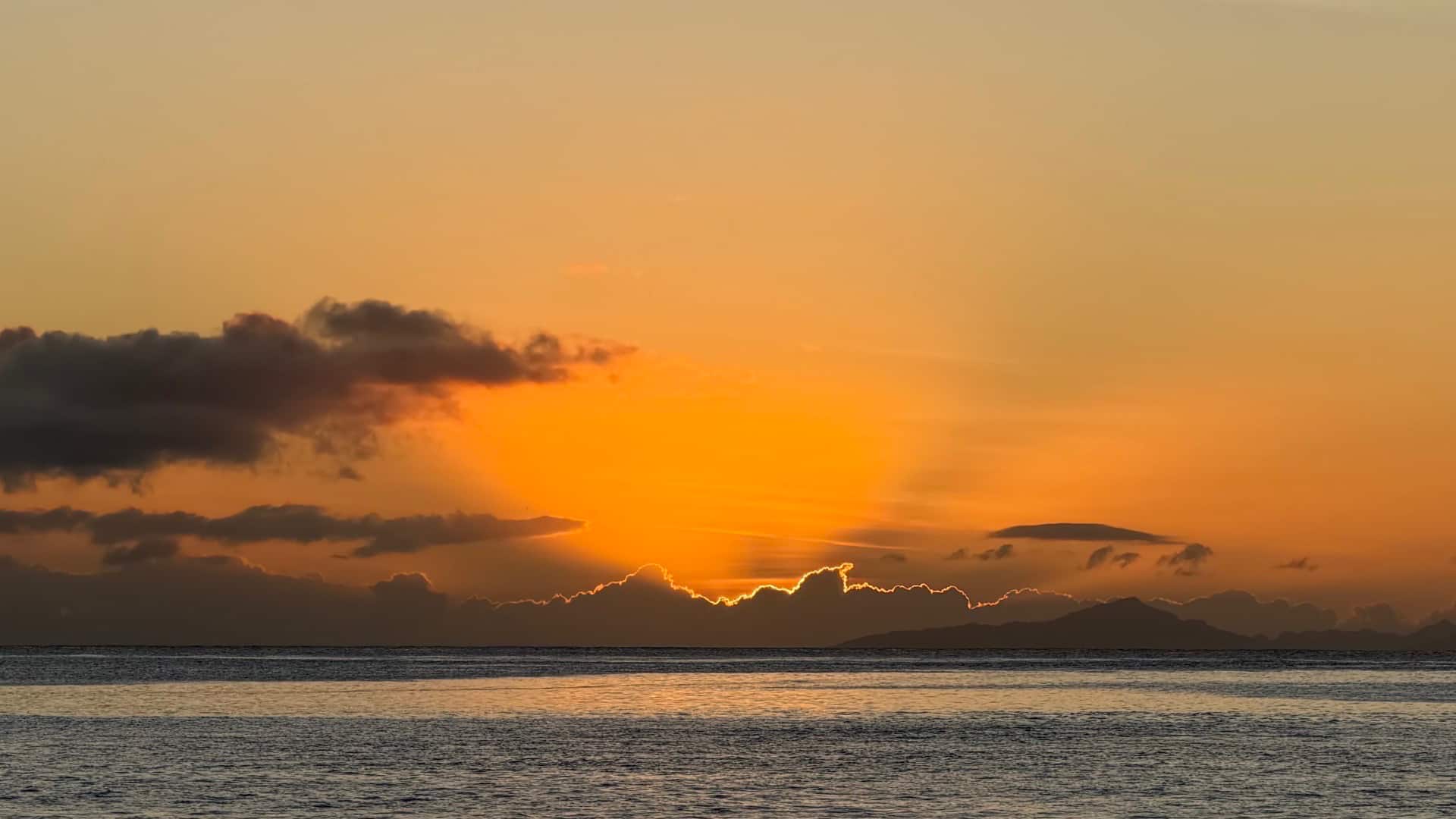 Vibrant golden sunset glowing behind dark ocean clouds over a calm sea horizon.