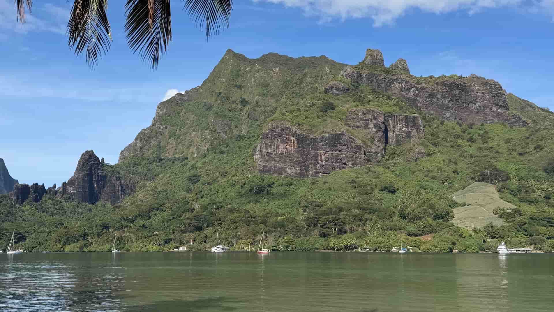Rugged dark basalt mountain cliffs rising above a calm coastal bay with anchored sailboats.