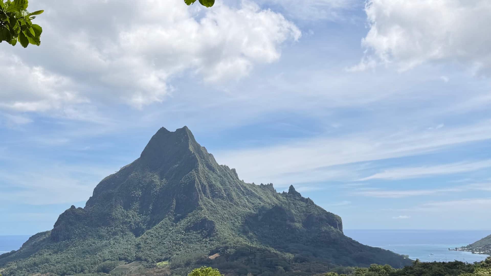 Majestic green volcanic mountain peak rising sharply against a partly cloudy tropical sky.