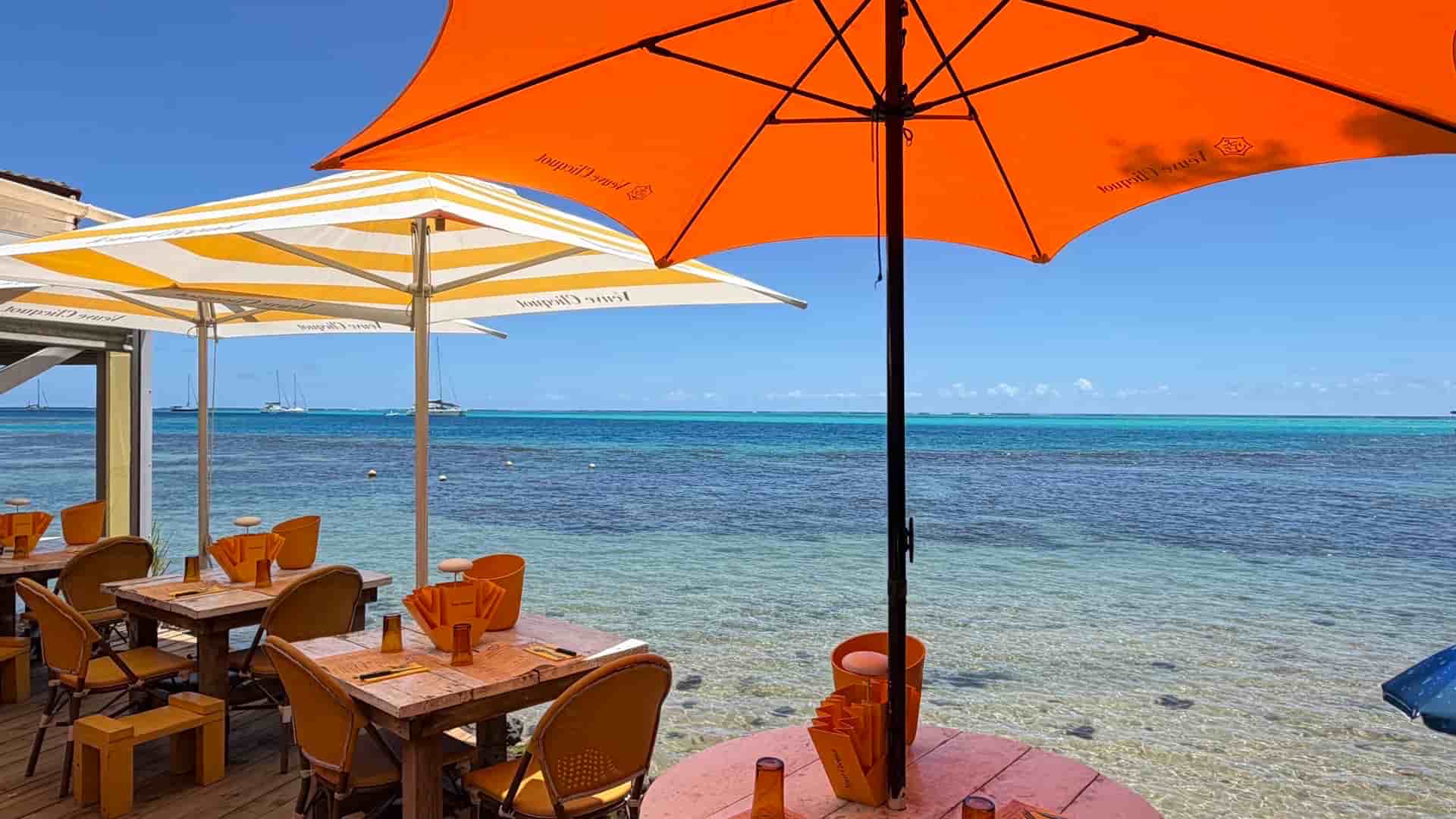 Orange and white parasols shading wooden dining tables on a sunny beachfront overlooking the turquoise lagoon.