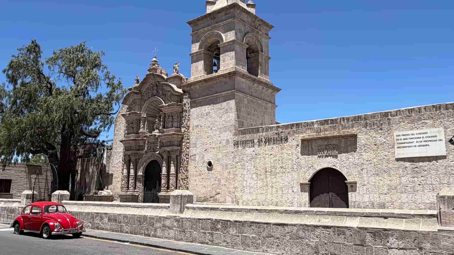 Vintage red Volkswagen Beetle parked along a historic sillar stone wall and church in Arequipa.