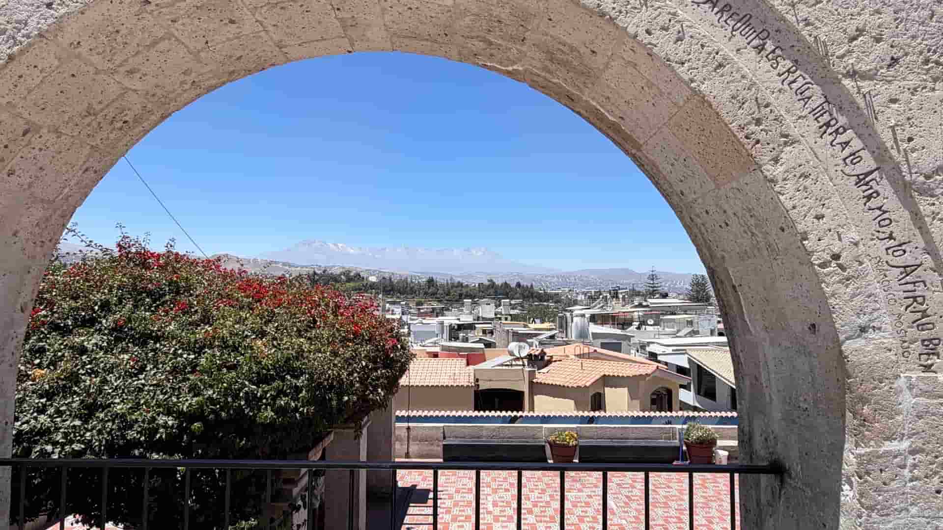 Panoramic view of Arequipa's terracotta rooftops and distant snow-capped volcanoes through a sillar archway.
