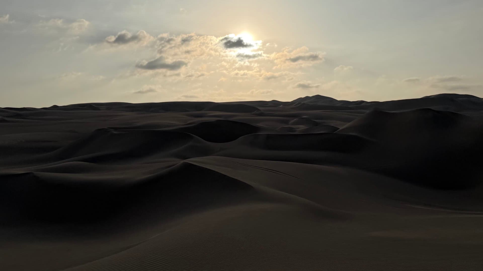 Golden sunlight breaking through scattered clouds over undulating desert sand dunes.