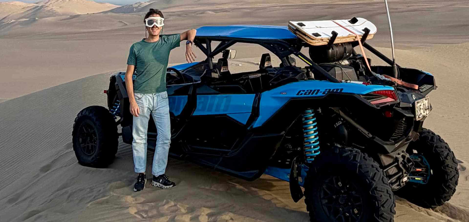 Traveler posing next to a blue Can-Am dune buggy parked on a sandy desert ridge.