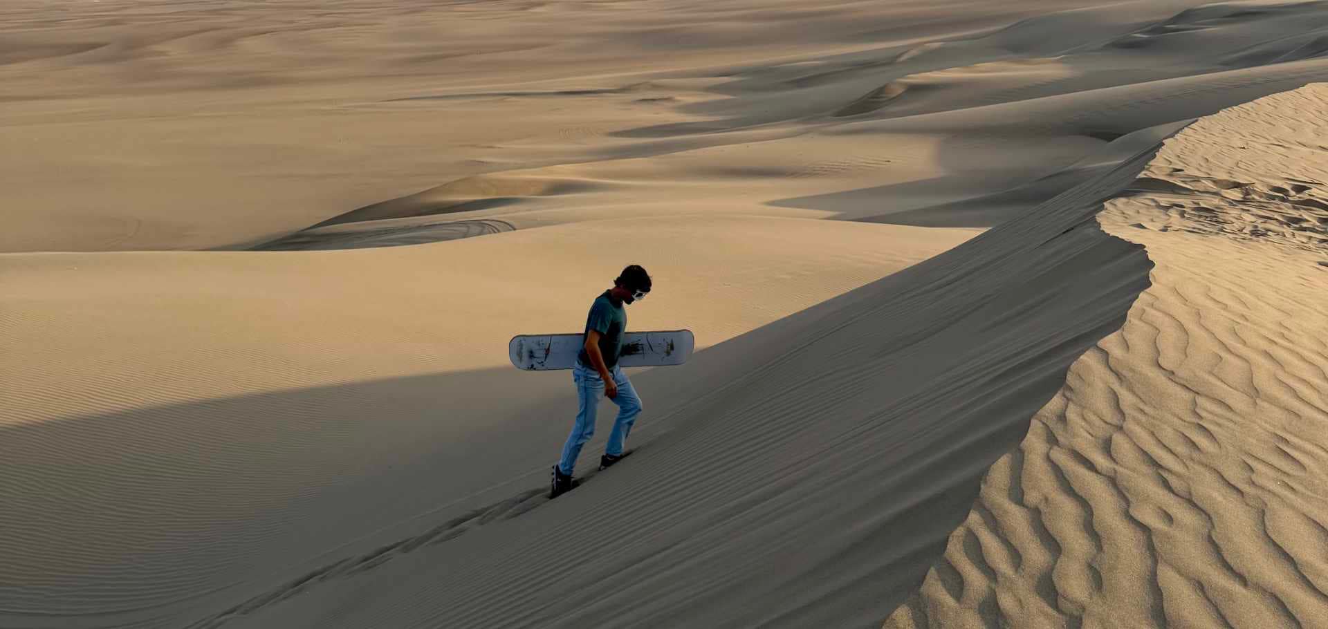Traveler carrying a sandboard while walking up the ridge of a massive desert dune.