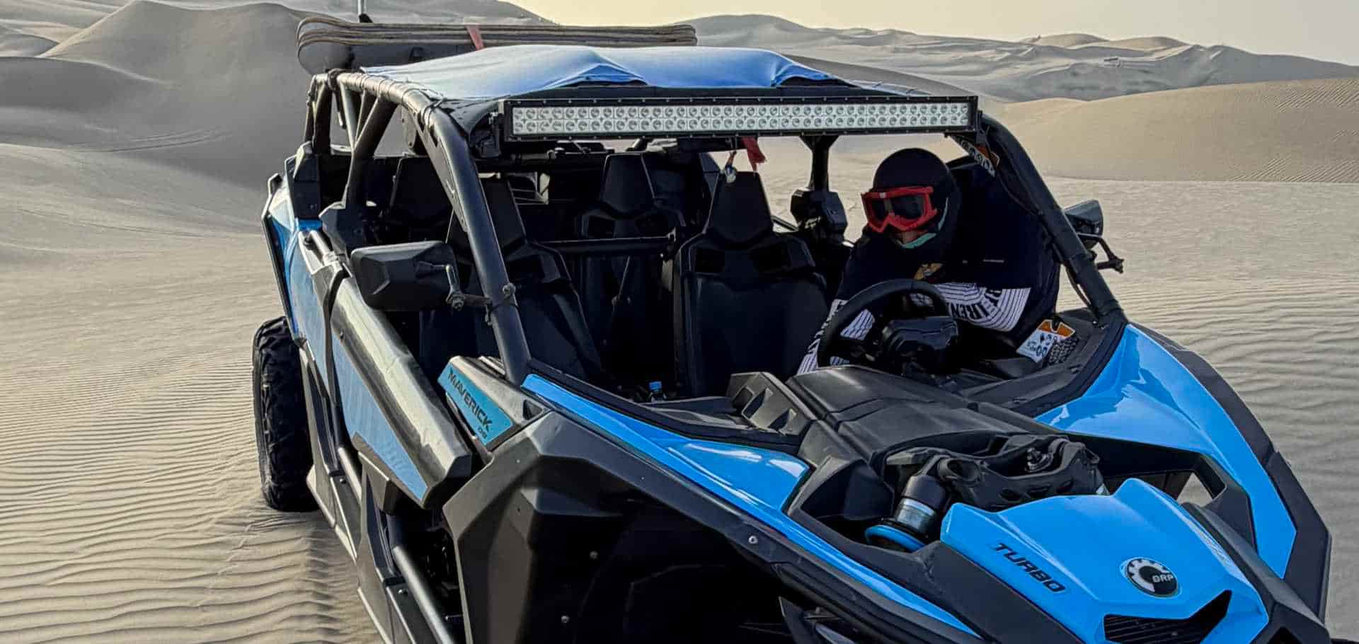 Blue Can-Am sand buggy parked on the desert dunes with a red safety flag.
