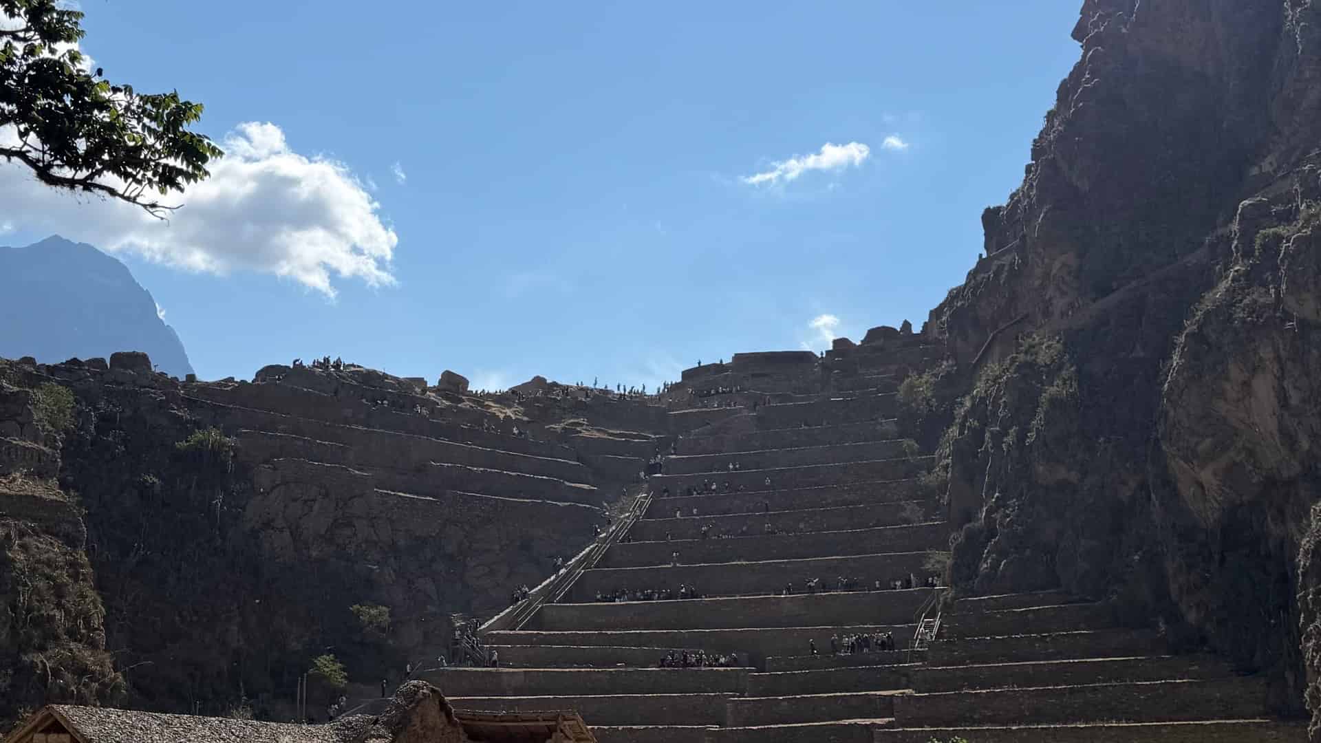 Tourists ascending the massive stone agricultural terraces under a bright blue sky.